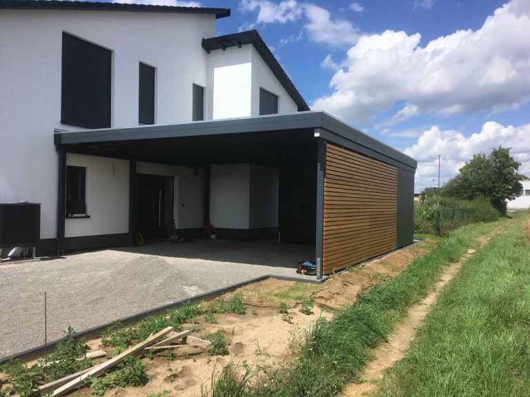 Graue Garage mit Holzverkleidung neben einem modernen Wohnhaus und grünem Wiesenbereich.