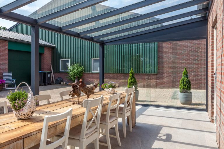 Holzterrasse mit Esstisch, Stühlen und Pflanzen unter einem transparenten Dach.